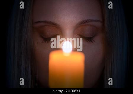 Nahaufnahme`s Gesichts einer Frau mit geschlossenen Augen im Dunkeln hinter einer verschwommenen Kerze mit Fokus auf den Hintergrund. Mystik, Glaube oder Religion Konzepte Stockfoto